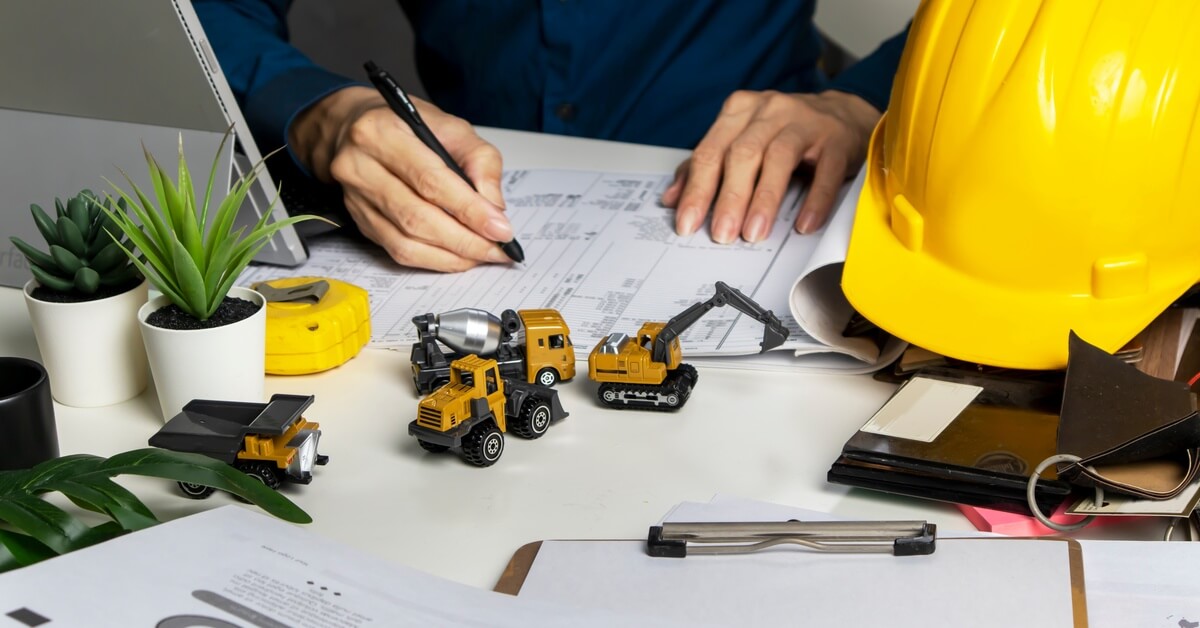 A person in a blue shirt writes on a printed chart at a desk covered in paperwork and miniature construction vehicles.