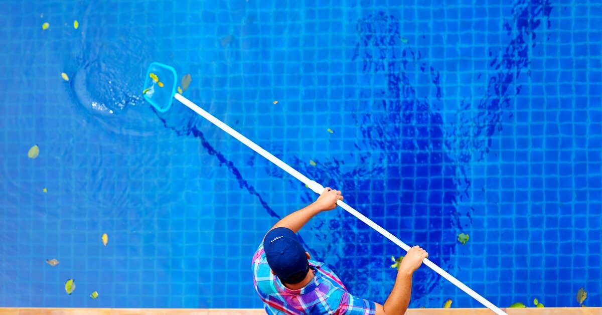 An aerial view of a young man wearing a hat and a plaid shirt who is using a cleaning pole to remove leaves from a pool.