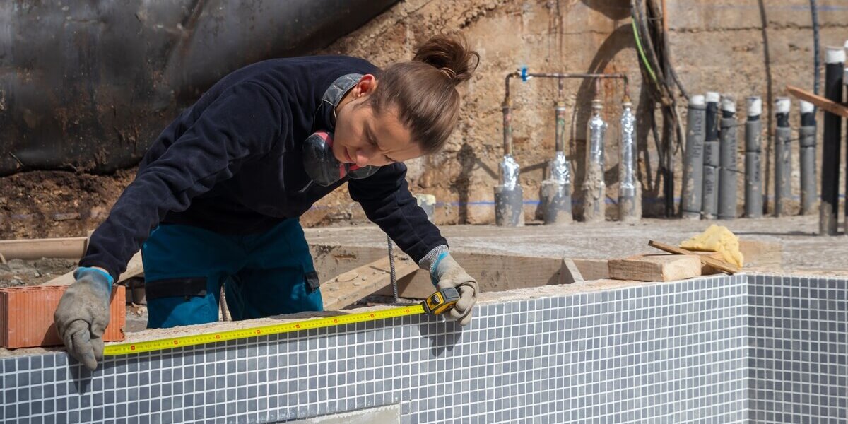 Construction worker using a measuring tape on the top of a pool during a renovation with pipes exposed in background.