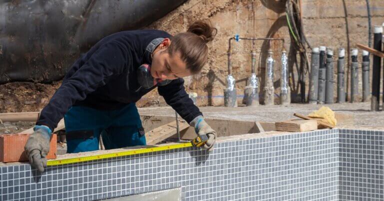 Construction worker using a measuring tape on the top of a pool during a renovation with pipes exposed in background.