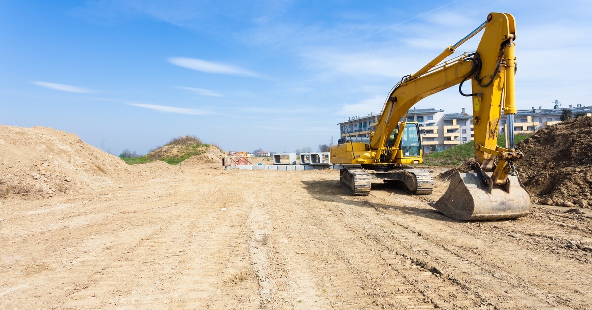 A construction site with a large plot of land cleared by a yellow excavator machine. Mounds of dirt surround the area.