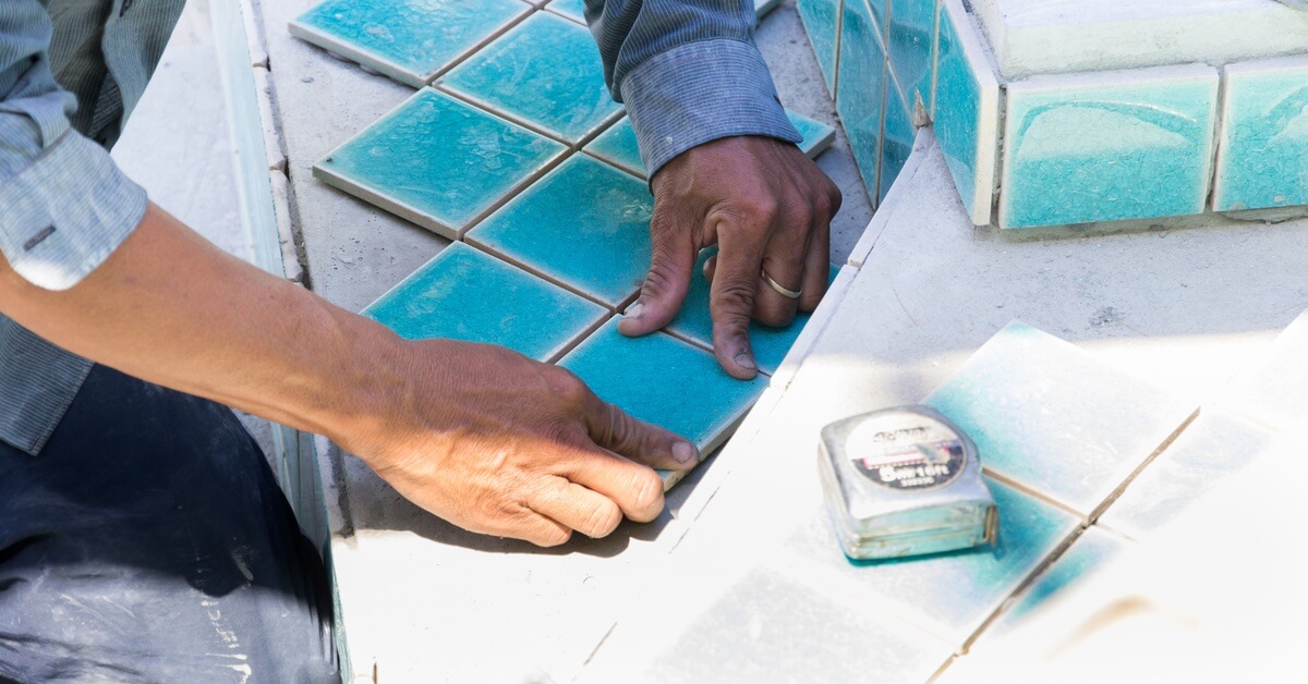 A close-up of a construction worker's hands installing blue square tiles on the steps of an empty pool.