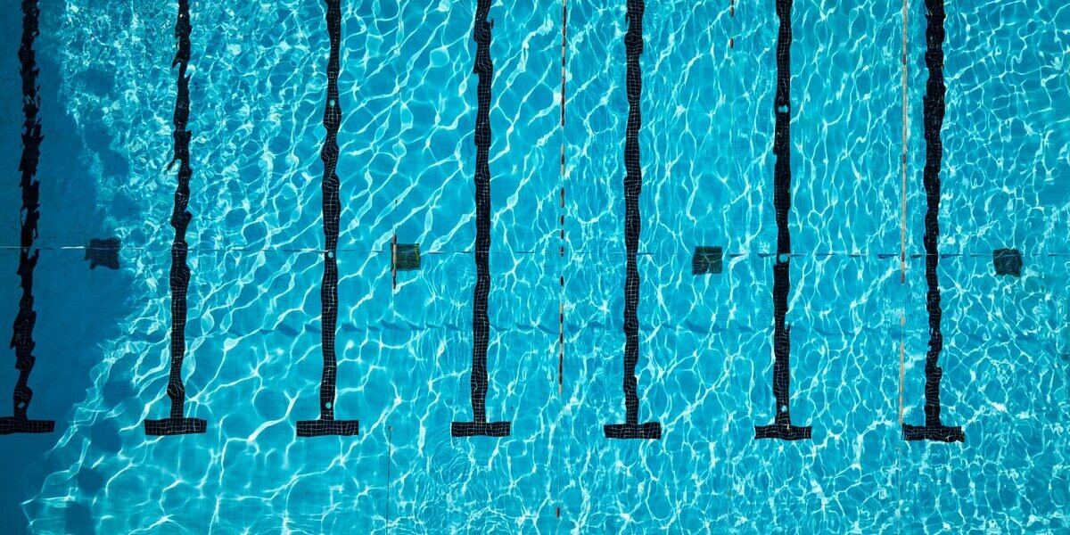 An aerial view of a large pool with clear water, straight black underwater lane lines, and small hanging flags.