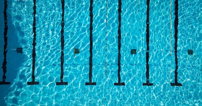 An aerial view of a large pool with clear water, straight black underwater lane lines, and small hanging flags.