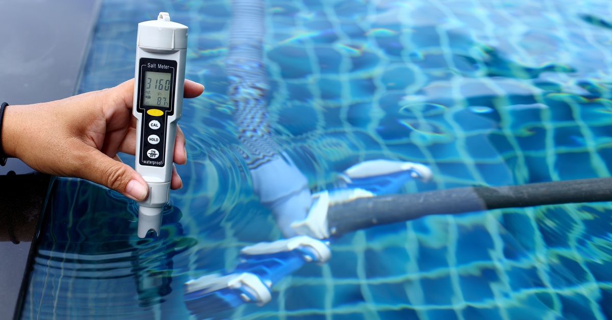 A hand, up close, holding a pool thermometer inside the water at the edge of a pool. Gray piping is underneath the water.