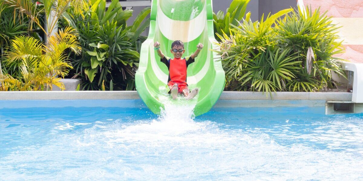 A kid wearing a full-body red bathing suit sliding down a green waterslide into a large pool with greenery in the background.
