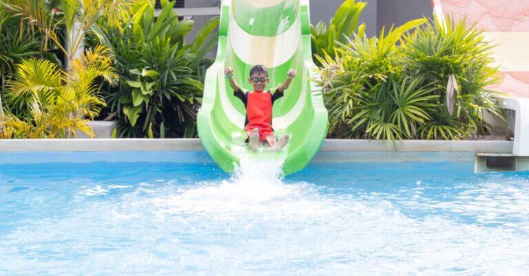 A kid wearing a full-body red bathing suit sliding down a green waterslide into a large pool with greenery in the background.