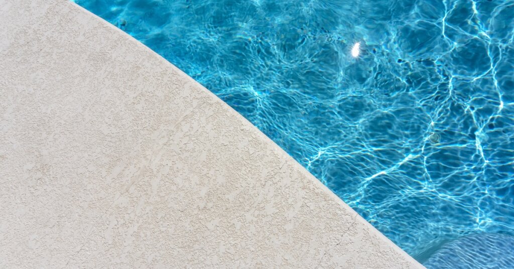 A close-up of the curved concrete edge of a pool filled with clean blue water reflecting ripples of sunlight.