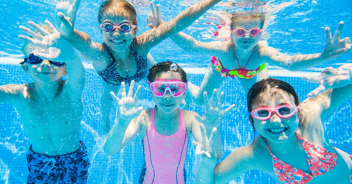 Multiple kids wearing bathing suits and goggles, smiling under crystal clear water in a pool on a sunny day.