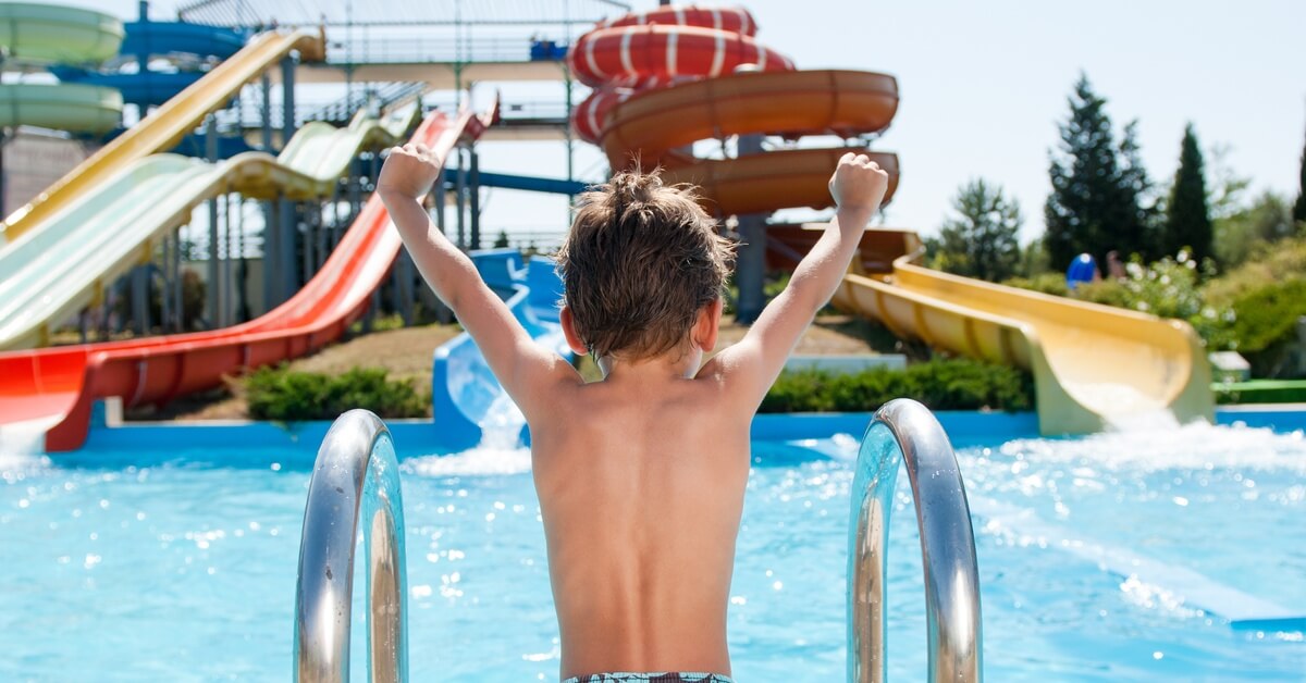 A boy raising both arms in front of a large pool with clear water and multiple waterslides in the background.